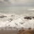 Small Sandy Hidden Beach with Rocks. Waves Washing Over the Sandy Beach. Sun is Lighting Up the Foreground. Cloudy Skies in Background.